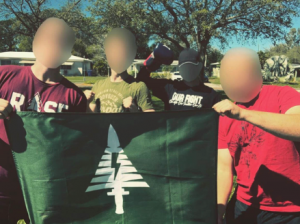 Four people stand outdoors in front of trees and recreational vehicles, holding a dark green banner. Their faces are digitally blurred for anonymity. From left to right: one person wears a maroon shirt with "ROSE" partially visible, another wears an olive green shirt, a third wears a black shirt with "Proud Boys" text visible on the sleeve, and the fourth wears a red shirt. The banner displays a white graphic combining a pine tree shape with an integrated lightning bolt running vertically through its center.