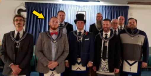 Group photo of eight men standing indoors in front of blue curtains, dressed in formal attire with Masonic regalia including decorative aprons and collars with symbolic emblems. A yellow arrow points to a man in the front row wearing a gray suit jacket and red bow tie. Center figure wears a black top hat and dark suit. Other members wear suits, turtlenecks, and various decorated aprons featuring geometric patterns and symbols. Framed pictures hang on the wall behind them. The men stand in two rows, with some in the back row partially obscured.