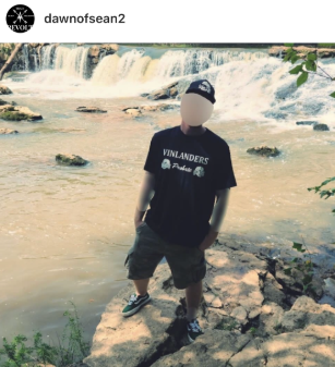 Instagram post by user "dawnofsean2" showing a person standing on a rocky bank in front of a wide, cascading waterfall. The person's face is obscured by a blank oval blur. They wear a black t-shirt printed "VINLANDERS" in white letters above a small logo, a black beanie with a white emblem, gray cargo shorts, and black-and-white sneakers. Turbulent pale-brown water flows over the falls behind them, with trees and foliage framing the upper edges of the frame. A circular profile avatar with a stylized design appears in the upper left corner.