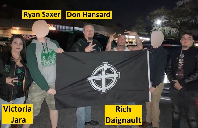 Night photo of six people standing outdoors in a parking lot holding a black flag with a white circular Celtic cross symbol. From left to right: Victoria Jara, a woman with dark hair in a black jacket with patches including "1488" visible, makes a peace sign with her right hand. Ryan Saxer, with his face digitally blurred, wears a gray-and-green raglan hoodie with a cyclist and tree graphic, making a gesture with his right hand. Don Hansard, an older man with short hair, gestures with both hands in front of his chest. Rich Daignault, wearing a dark tank top, raises both hands in a gesture near his head. Two additional people with digitally blurred faces stand on the right, one in a dark jacket and another in a dark shirt with text visible. Parked vehicles, streetlights, and trees appear in the background under nighttime lighting. Yellow label boxes identify four individuals by name above or below them.