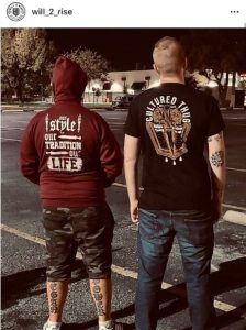 Screenshot of an Instagram post by user "will_2_rise" depicting two men standing in a parking lot with their backs to the camera. The man on the left wears a maroon hoodie with white text reading "I speak ONLY THE TRUTH 313" and camouflage shorts, revealing tattoos on his calves. The man on the right wears a black t-shirt with a large winged graphic and text on the back, blue jeans, and a tattoo on his left arm. Trees and commercial buildings are visible in the background under bright sunlight.