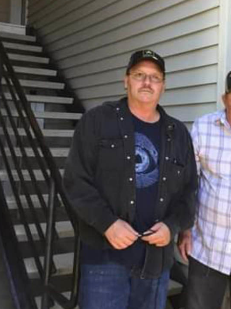 Photo of a middle-aged man with glasses and a mustache standing outdoors in front of light-colored horizontal siding. He wears a dark baseball cap, a dark button-up overshirt over a navy t-shirt with a blue circular eye graphic, and blue jeans. His hands hold what appears to be eyeglasses in front of him. Black metal stairs with railings visible behind him to the left. Partial view of another person in a light plaid shirt on the right edge.