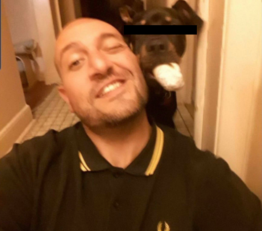 A selfie of Vito Russel, a bald man with light beard stubble, smiling broadly, wearing a black polo shirt with gold collar trim and a small logo. Behind him in a hallway, a dark-colored dog holds a white object in its mouth; the dog's eyes are covered with a black censor bar.